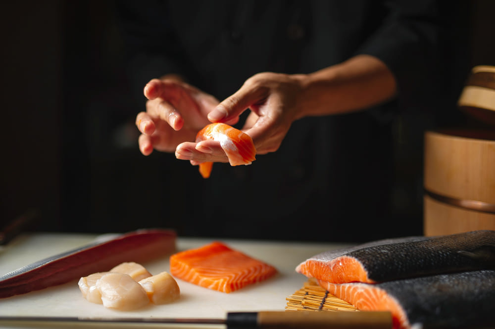 A culinária japonesa já faz parte do gosto dos brasileiros. Claro, aqui os tradicionais pratos do país aparecem com uma pitadinha de Brasil, mas sempre com as raízes orientais presentes. Os sushis são um ótimo exemplo disso, e aqui em nosso país, vemos até nas versões doces. Mas, afinal, como esse prato típico é consumido no Japão? Quais são os peixes para sushi mais comuns na Terra do Sol Nascente? Aqui no Brasil, estamos habituados a usar o salmão como principal peixe de sushi. Curiosamente, essa espécie não fazia parte da culinária japonesa até os anos 1980, quando os noruegueses o apresentaram para o Japão. No entanto, o país sempre teve uma incrível variedade de peixes e frutos do mar, que muitas vezes são desconhecidos pelos estrangeiros que vão conhecer a gastronomia local. Alguns deles já são conhecidos por aqui, outros, não. Confira! Atum O atum é mais um peixe que caiu no gosto dos ocidentais. Conhecido pelo seu sabor intenso, teor de gordura moderado e consistência firme, é o tipo de peixe para sushi que agrada pela sua intensidade e sabor. Pargo No Japão, o pargo é um peixe especial, consumido em ocasiões festivas. No Ano Novo, ele é normalmente assado inteiro e consumido. Mas também é um peixe utilizado em sushis, trazendo um sabor delicado e doce para o prato. Ovas de salmão As ovas de peixe para sushi são comumente utilizadas na gastronomia local, reconhecidas por explodirem na boca. Elas têm uma textura macia e coloração vermelha brilhante. As mais utilizadas na culinária são: Massago (capelim), Tobiko (voador) e a Ikura (salmão). Polvo No quesito frutos do mar, o polvo pode ser utilizado para fazer sushis cheios de sabores intensos. Além desse prato, o alimento também pode ser usado nos tradicionais takoyaki, que são bolinhos de polvo. Lula O sushi de lula é uma outra opção consumida no Japão e que também já caiu no gosto dos brasileiros. Com textura cremosa e macia, é uma ótima aposta para vegetarianos. Como fazer os cortes dos peixes? Um dos maiores desafios na hora de preparar essa iguaria é saber como cortar peixe para sushi. O principal segredo está na lâmina, que deve ser fina, longa e bem afiada. A tábua pode ser de madeira ou plástico, mas cuidado com as de vidro: elas costumam escorregar quando você trabalha com o peixe. Em um primeiro momento, tire o excesso de umidade do filé, depois verifique se existem espinhas na peça. Também é preciso descolar a gordura da carne, além de tirar a pele do peixe. Esses são os passos iniciais, mas os cortes especificamente podem variar de acordo com o tipo de sushi que você vai fazer. Deu vontade de sushi? Pede no Hirota! Deu para perceber que existe uma variedade grande de peixe para fazer sushi, não é mesmo? Seja você da turma que pede no delivery ou então que prefere cozinhar, no Hirota existem inúmeras opções para matar a vontade desse prato delicioso. No Hirota Food Express, temos vários combinados de sushi, além de temakis e outros pratos orientais. Já no Hirota, você encontrará os ingredientes perfeitos para preparar sushis e os mais variados tipos de comidas. Conheça!